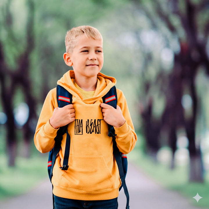 Young boy wearing a yellow hoodie with 'The Beast' text, holding a backpack in a park setting.