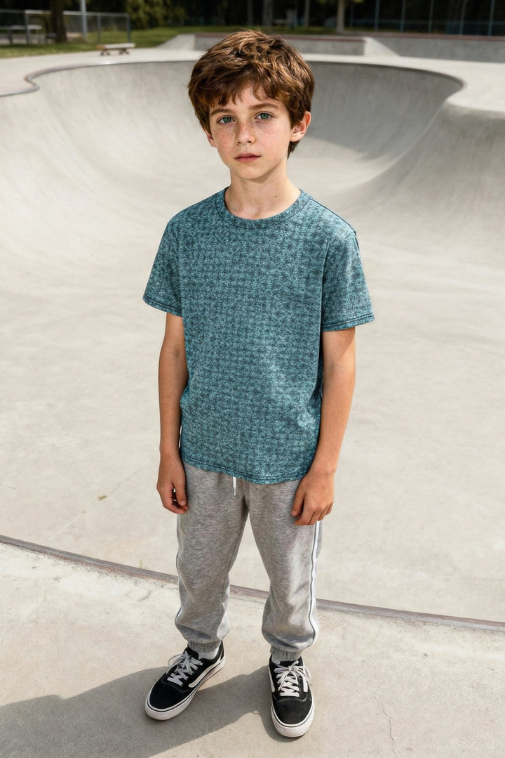 Young boy standing in a skate park wearing a blue shirt and gray pants.