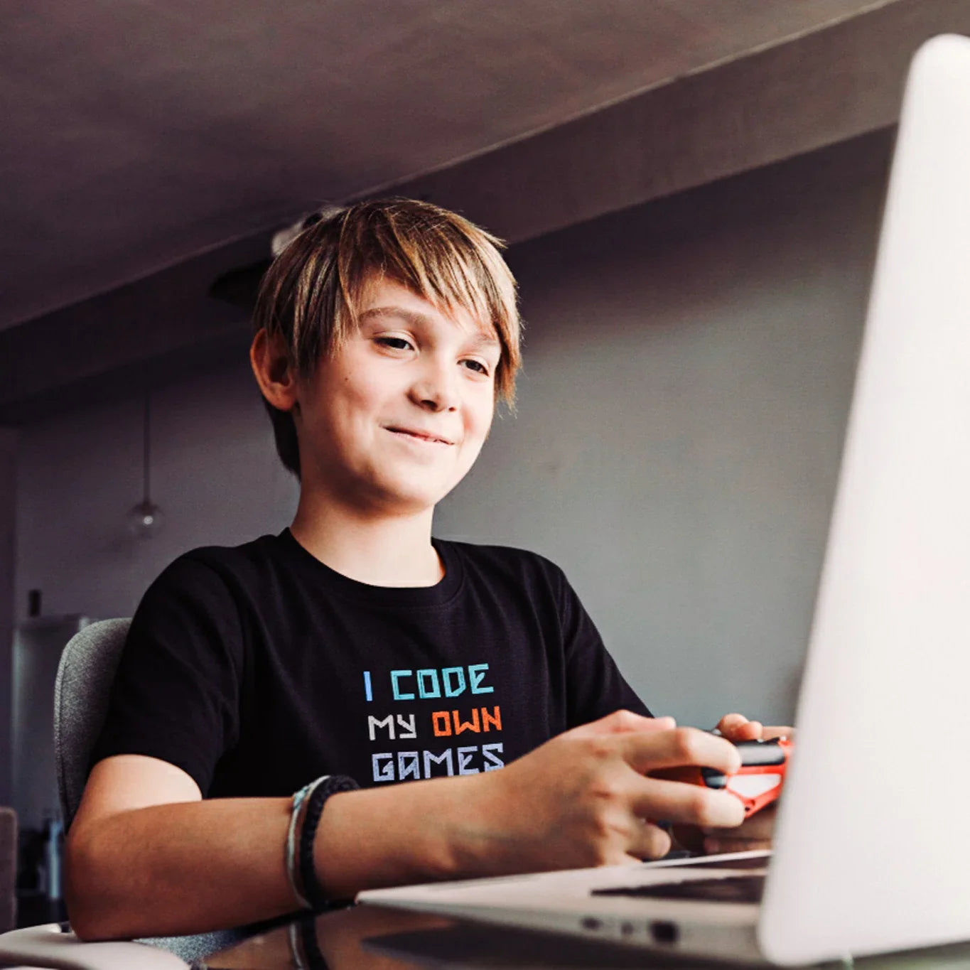 Child sitting at a desk with a laptop, wearing a black "I code my own games" House Panther t-shirt with text.