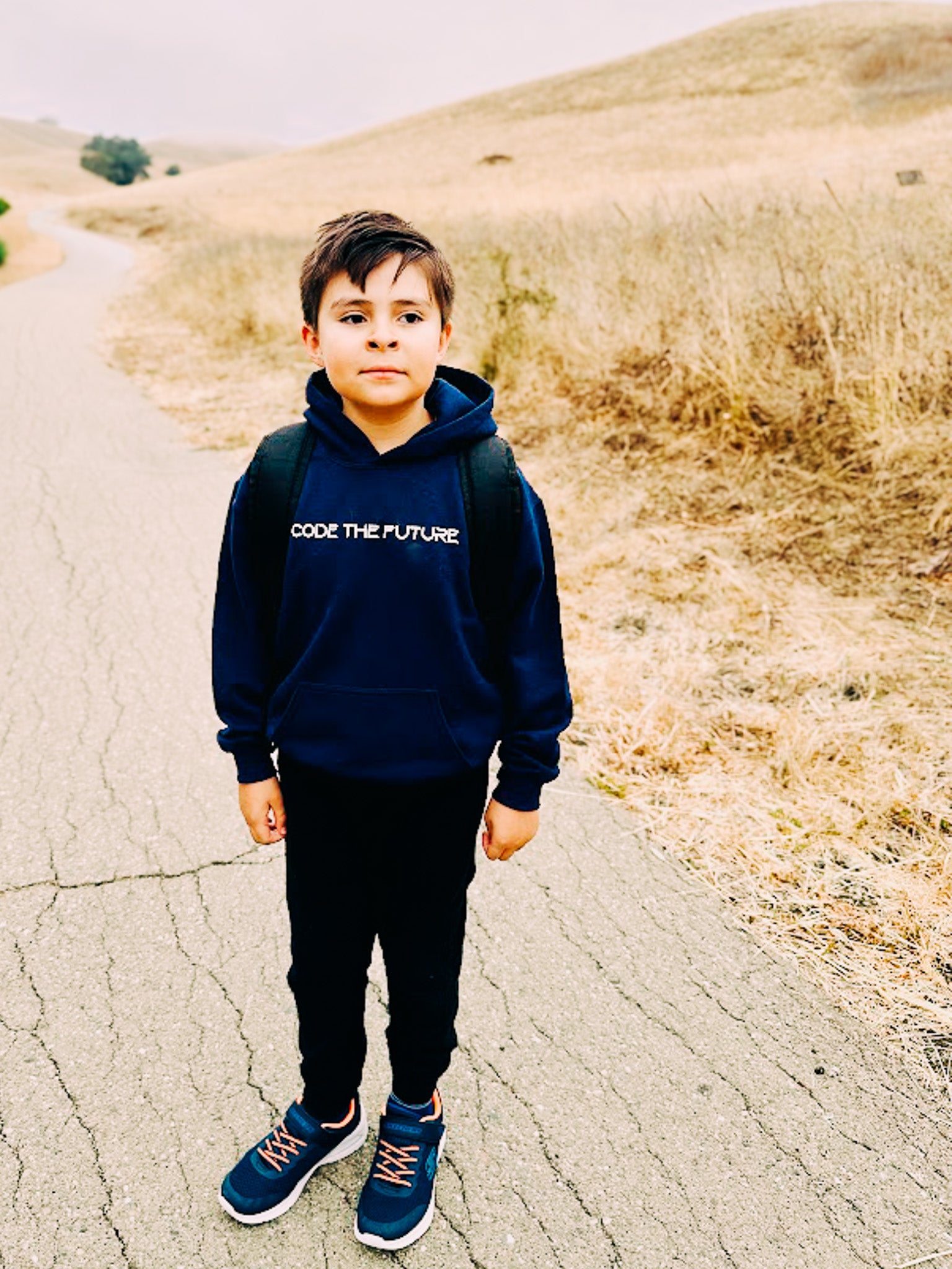 Child wearing a hoodie with text standing on a road with dry grass and hills in the background