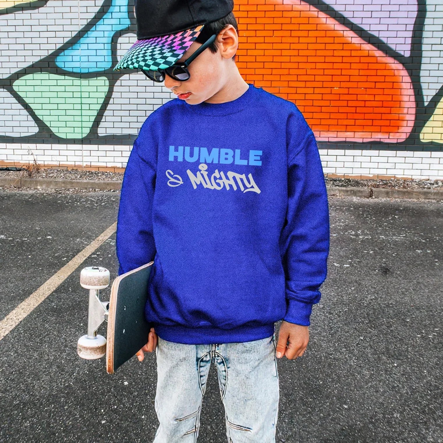 Child wearing a blue sweatshirt with 'Humble & Mighty' text, holding a skateboard in front of a colorful mural.