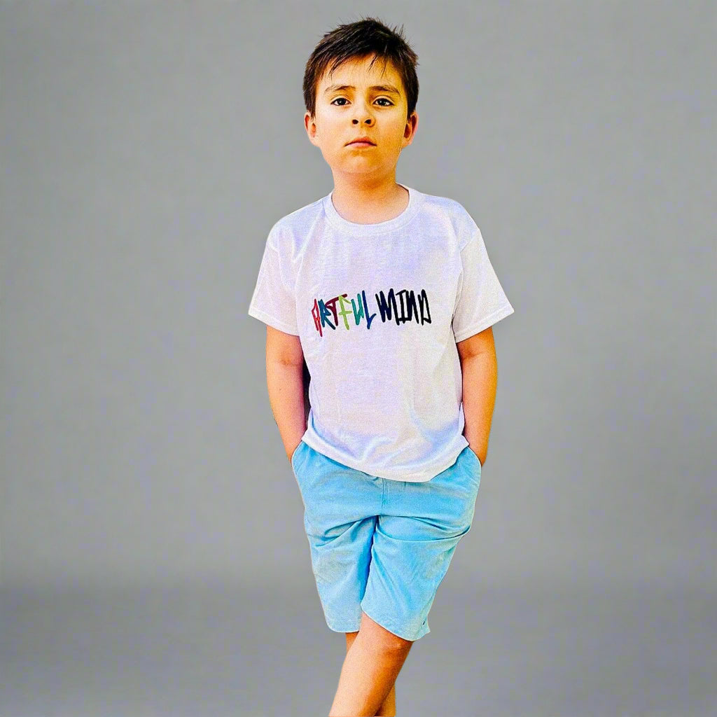 Child wearing a white t-shirt with colorful text and light blue shorts, standing against a beige wall.