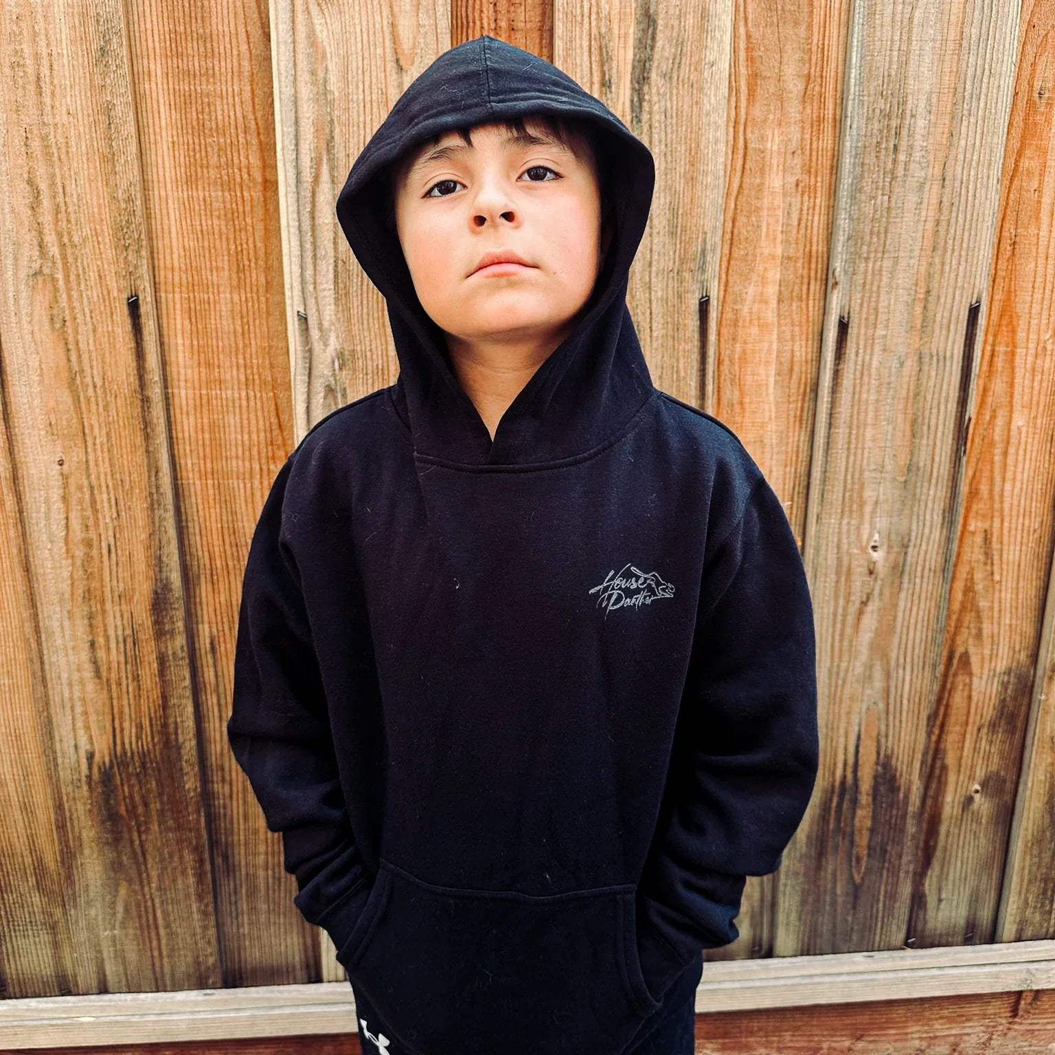 Child wearing a black hoodie with a logo against a wooden fence background