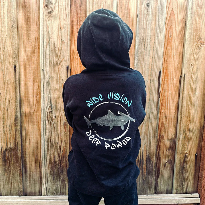 Person wearing a black hoodie with a logo and text, standing against a wooden fence.