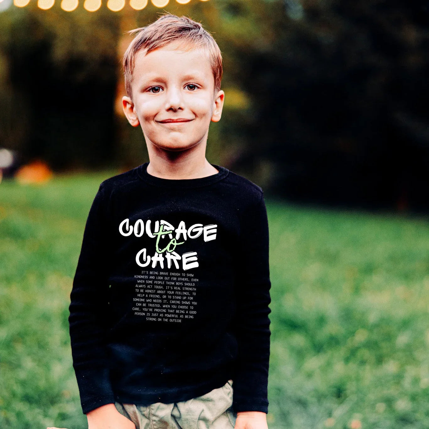 Child wearing a black sweatshirt with text, in a grassy outdoor setting with lights in the background.